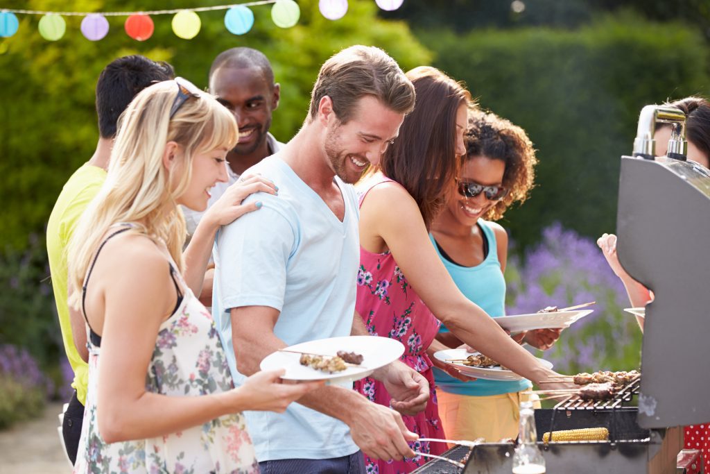 Smiling people standing around a grill, holding food or working on the grill.