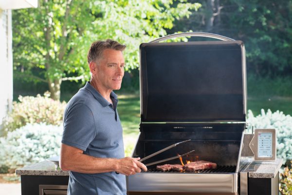 An image of a man wearing a blue shirt, standing by a grill with metal tongs in his hand.