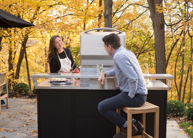 a woman grilling and talking to a man who is seated in front of a metal table